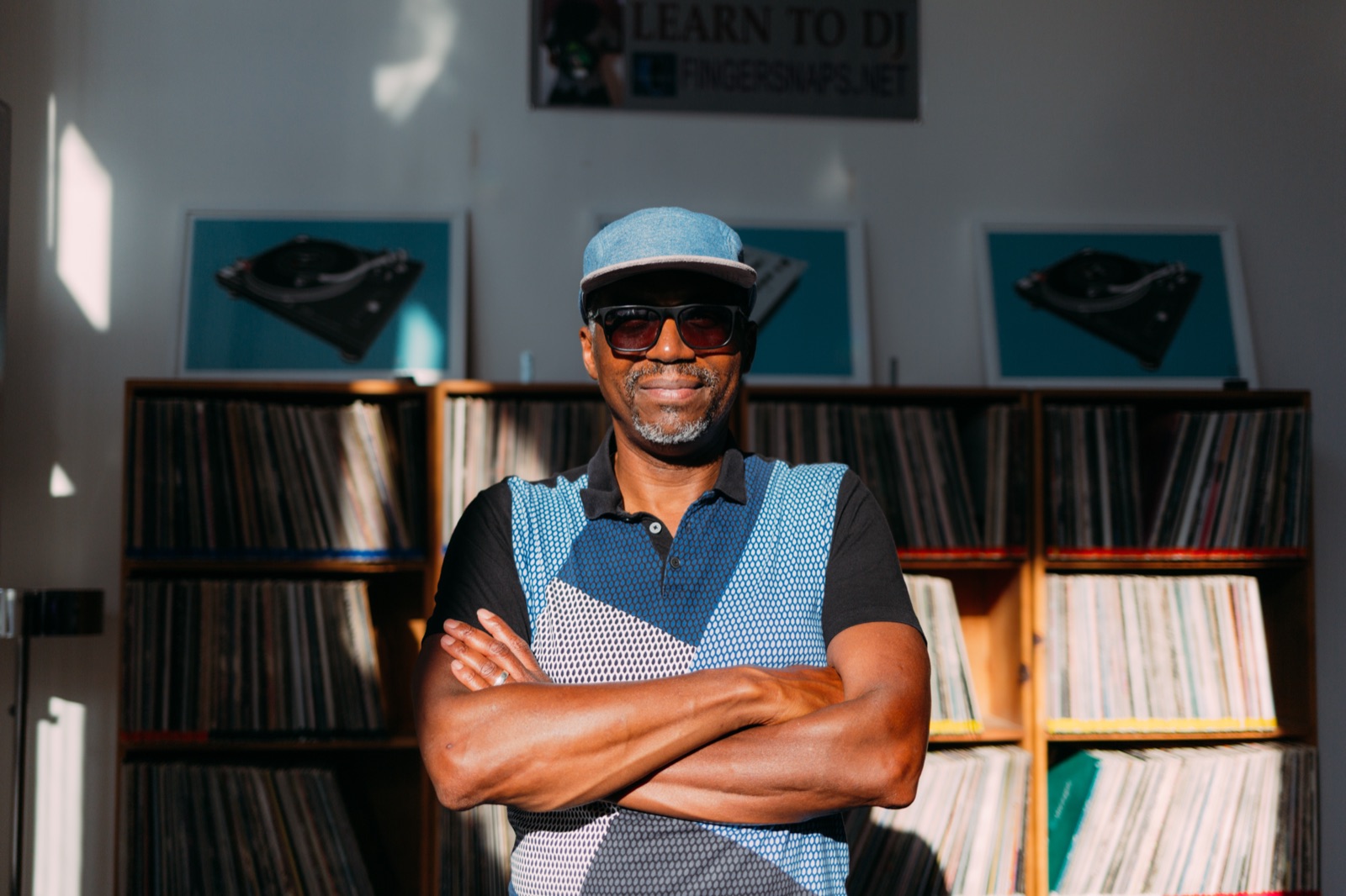 A record shop owner stands with his arms crossed in front of shelves of vinyl, under a sign reading "Learn to DJ".