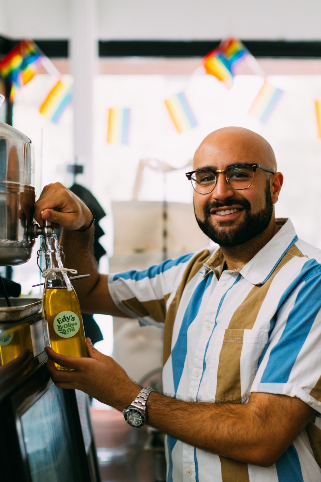 A grocer in glasses smiles at the camera while filling a glass bottle of olive oil from a bulk dispenser, Pride bunting strung above the shop window behind him.
