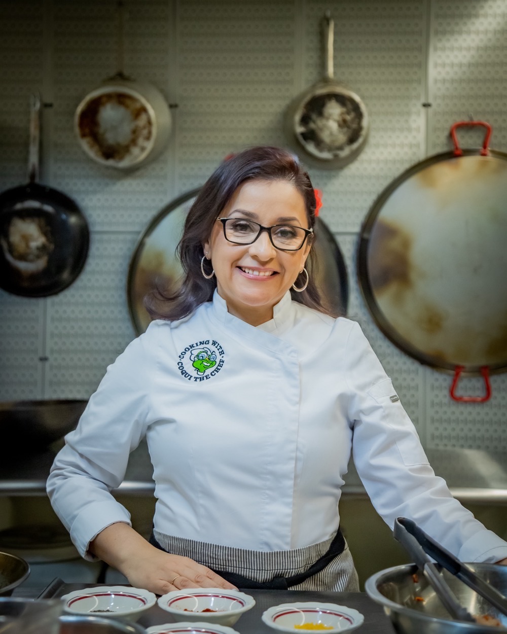 Tania Lopez of Coqui the Chef smiles at the camera in her chef's coat, standing at a stainless steel counter of prepped spices in a commercial kitchen hung with paella pans.