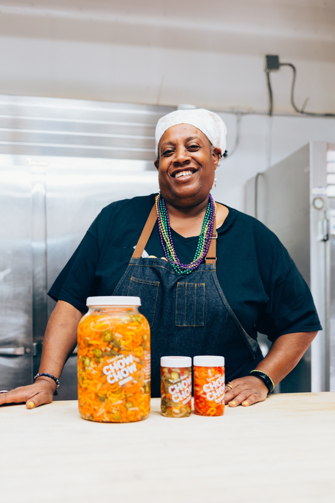 A food entrepreneur in a headscarf and colorful beads beams at the camera, her hands resting beside jars of her chow chow relish in her kitchen.