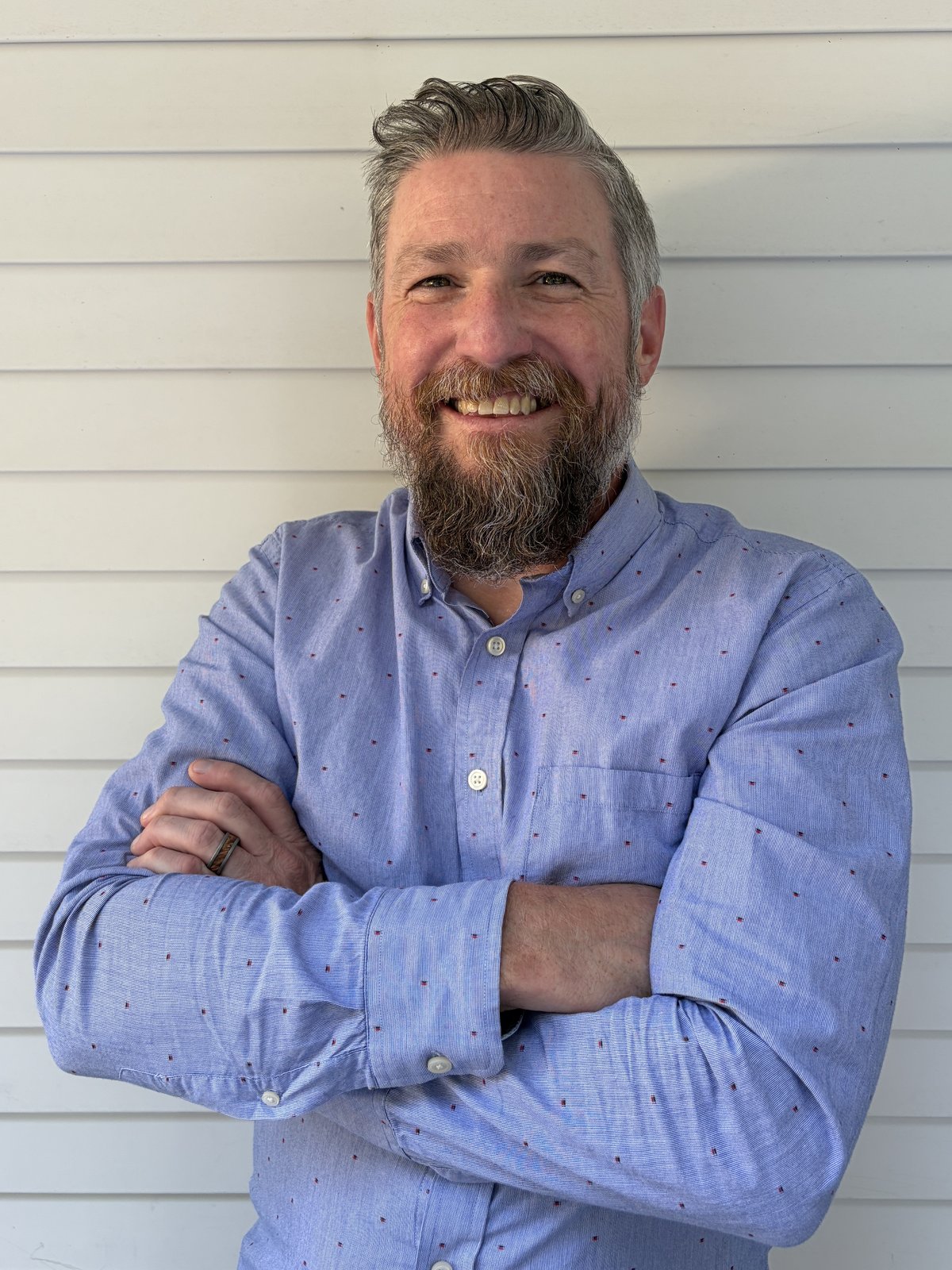 Brendan Landry, Start Small Think Big's Director of Corporate Engagement, arms folded and smiling in a light blue button-down shirt against white clapboard siding.