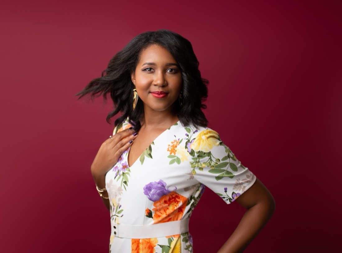 Elizabeth Lewis, founder of Brand, Style & Bloom, standing with one hand on her hip against a deep red backdrop in a white floral-print dress.