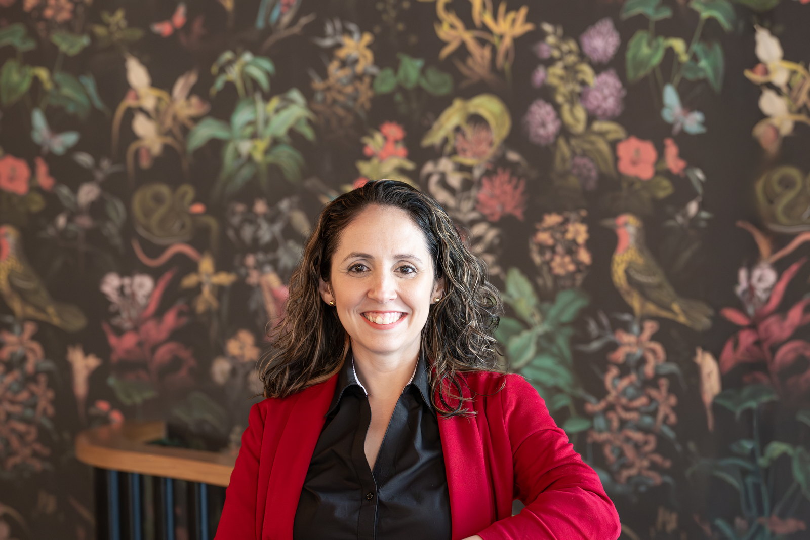 Mabell Fernandez, Start Small Think Big's Executive Director, smiling in a red cardigan and black shirt in front of a dark floral mural.