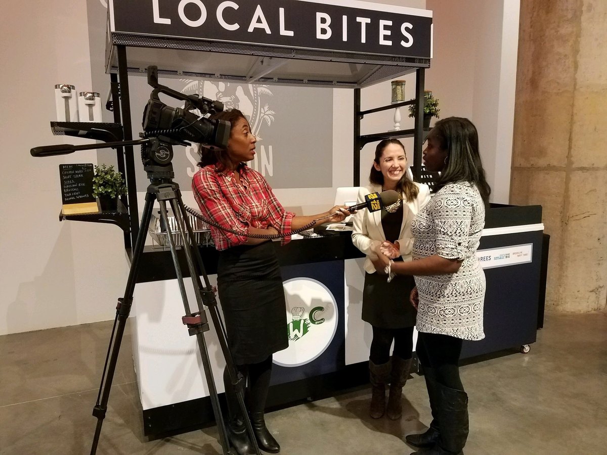 Mabell Fernandez of Start Small Think Big standing beside a woman speaking into a News 12 reporter's microphone, a television camera on a tripod alongside them, in front of the Local Bites food kiosk at the Brooklyn Navy Yard.