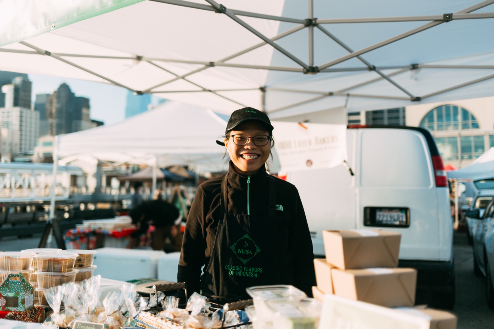 A smiling vendor in a Nusa cap and apron stands behind her farmers-market stall of baked goods, white canopy tents and the city skyline behind her.