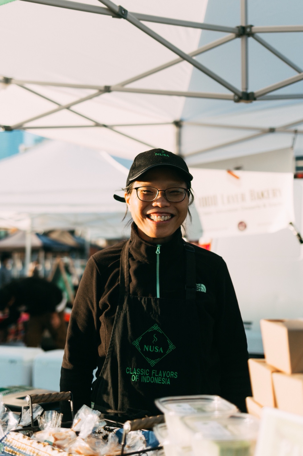 An Indonesian food business owner smiling behind her farmers market stand