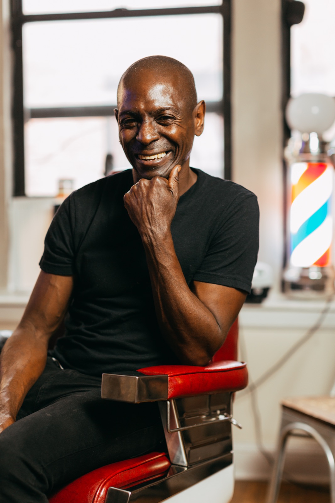 A barbershop owner laughs with his chin resting on his hand, seated in the red leather chair of his shop beside a glowing barber pole.