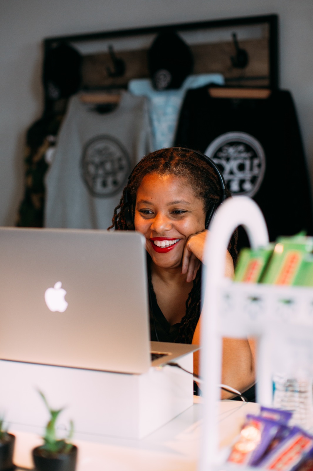 A small business owner wearing headphones smiles at her laptop in her spin studio, with branded apparel hanging on the wall behind her.