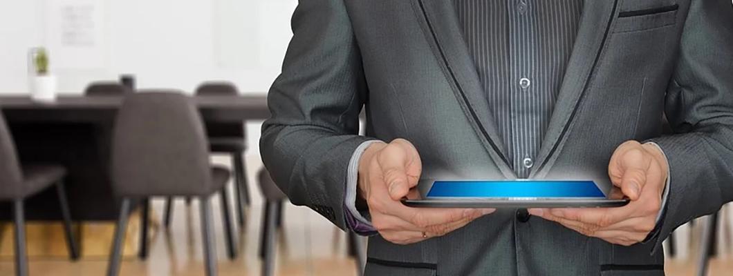 A man in a black suit in an office holding a tablet.
